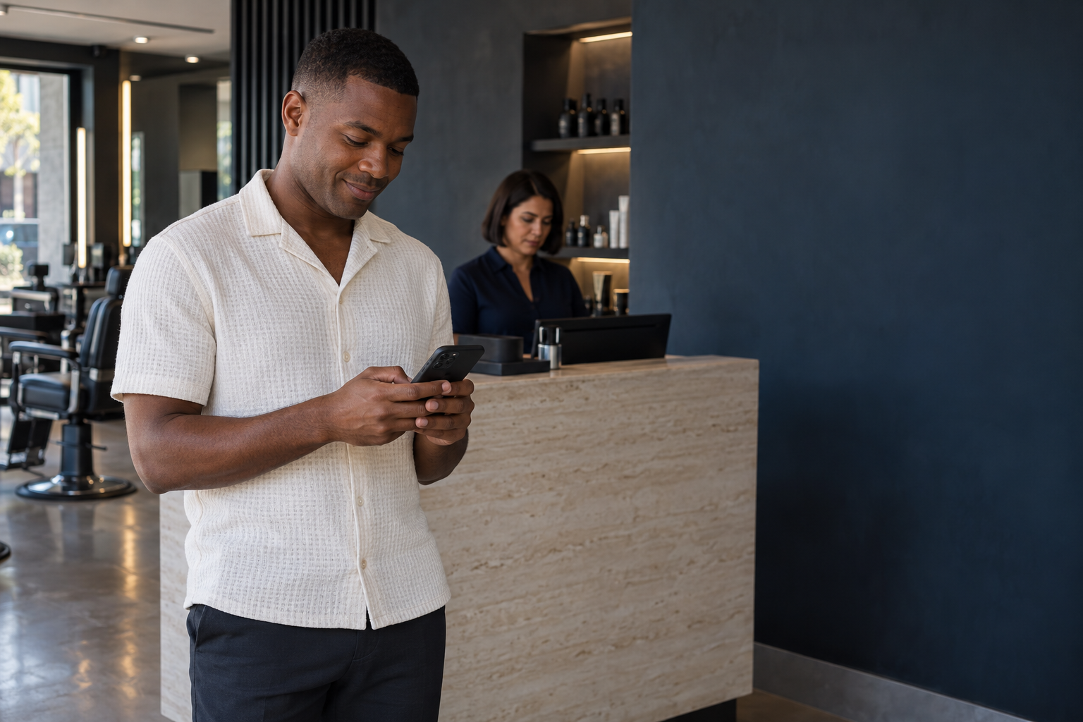 Cliente usando o celular na recepção de uma barbearia contemporânea, com uma gestora ao fundo.